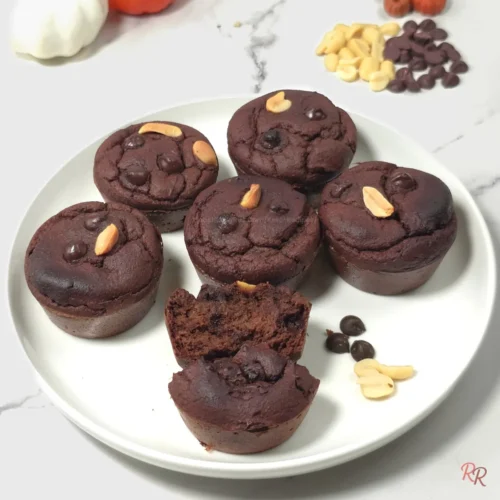 White plate of six keto chocolate muffins with peanuts and one muffin cut open to show rich interior.