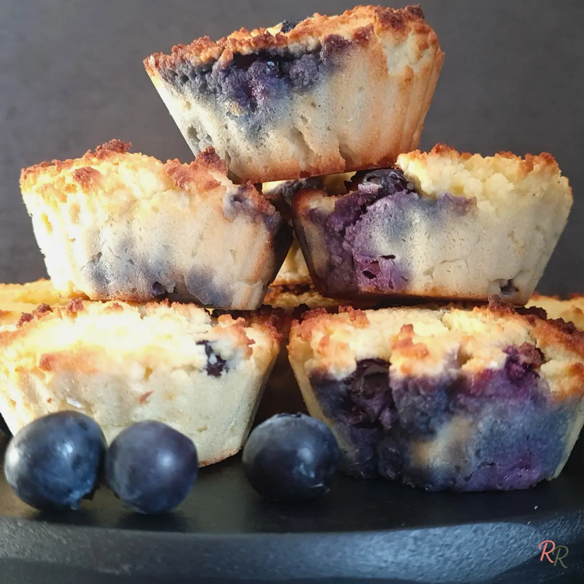 Close-up of keto blueberry muffins with browned coconut flour tops and juicy blueberries, stacked on a black plate.