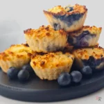 Stack of golden keto blueberry muffins made with coconut flour, served on a black plate with fresh blueberries.