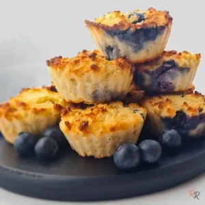 Stack of golden keto blueberry muffins made with coconut flour, served on a black plate with fresh blueberries.