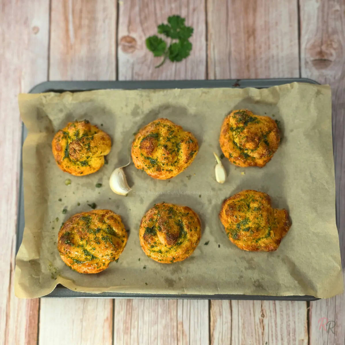 Overhead shot of six keto garlic knots on a parchment-lined baking tray, topped with parsley and garlic butter.