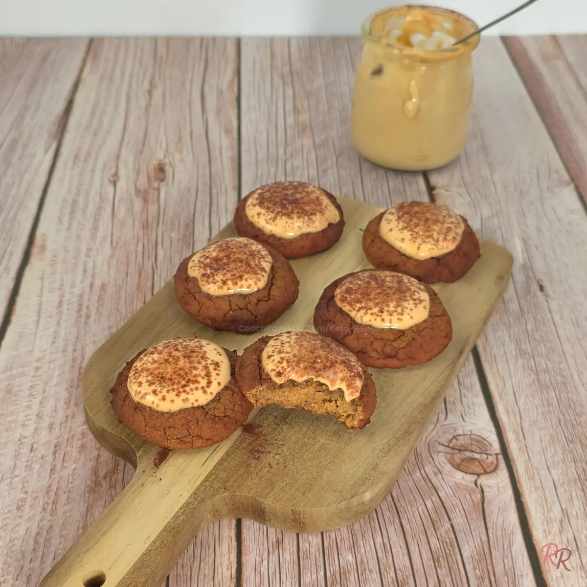 Six keto cottage cheese tiramisu cookies on a wooden board, topped with mousse and cocoa powder, with a jar of tiramisu mousse in the background.