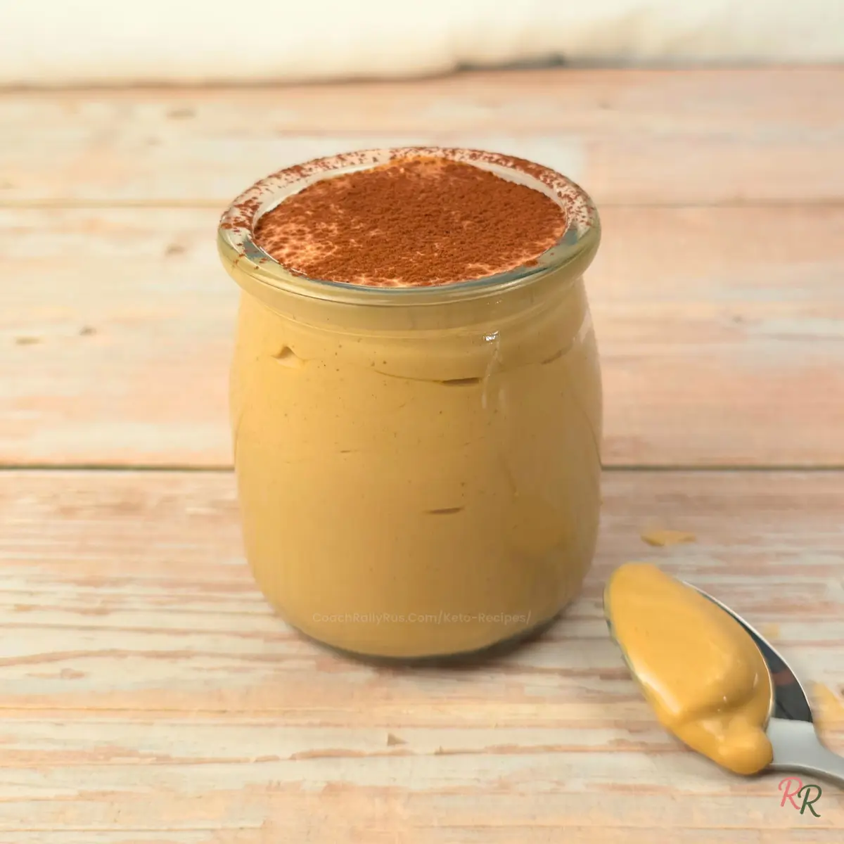 Side view of a glass jar filled with keto cottage cheese tiramisu mousse topped with cocoa powder on a light wood background.