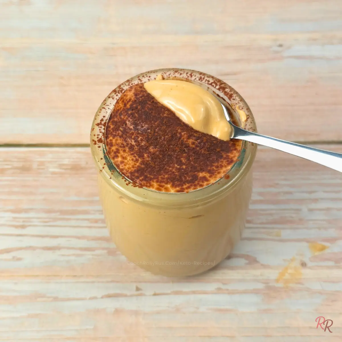 Close up of keto cottage cheese tiramisu mousse in a small glass jar with a spoonful lifted to show the smooth, creamy texture and cocoa powder dusting on top.