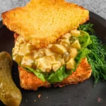 Creamy dill pickle cottage cheese egg salad served on toasted bread with lettuce, fresh dill, and a whole pickle on a black plate.