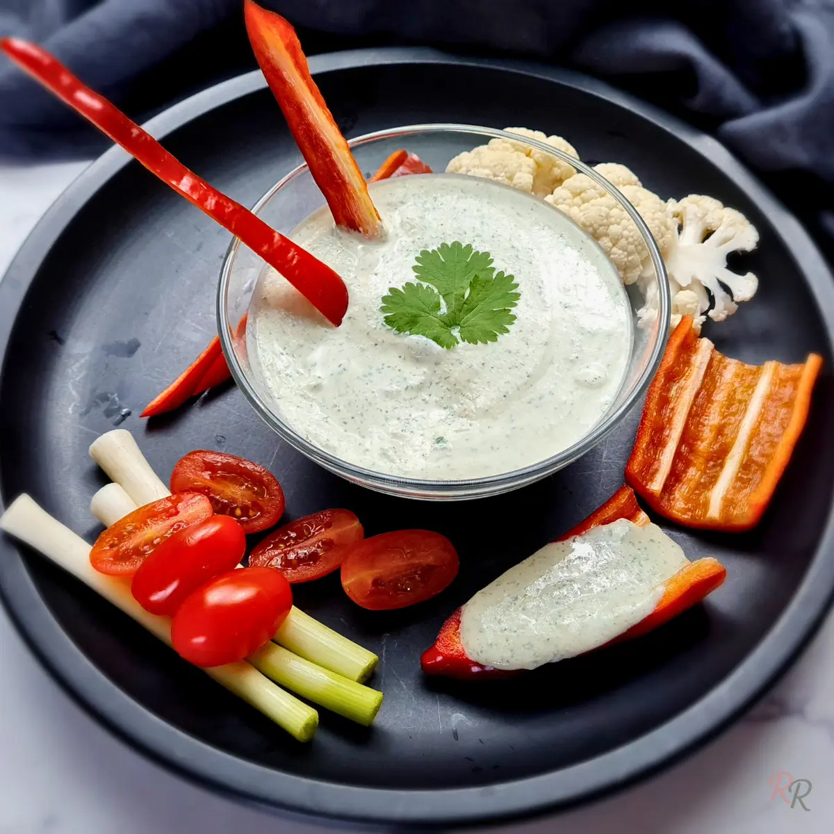 Keto Cottage Cheese Ranch Dip with Vegetables Glass bowl of keto cottage cheese ranch dip served with celery, cherry tomatoes, cauliflower, and red pepper on a black plate