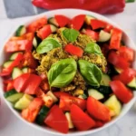 Keto chicken salad with pesto chicken, chopped tomatoes, cucumber, red bell pepper, and basil leaves in a white bowl