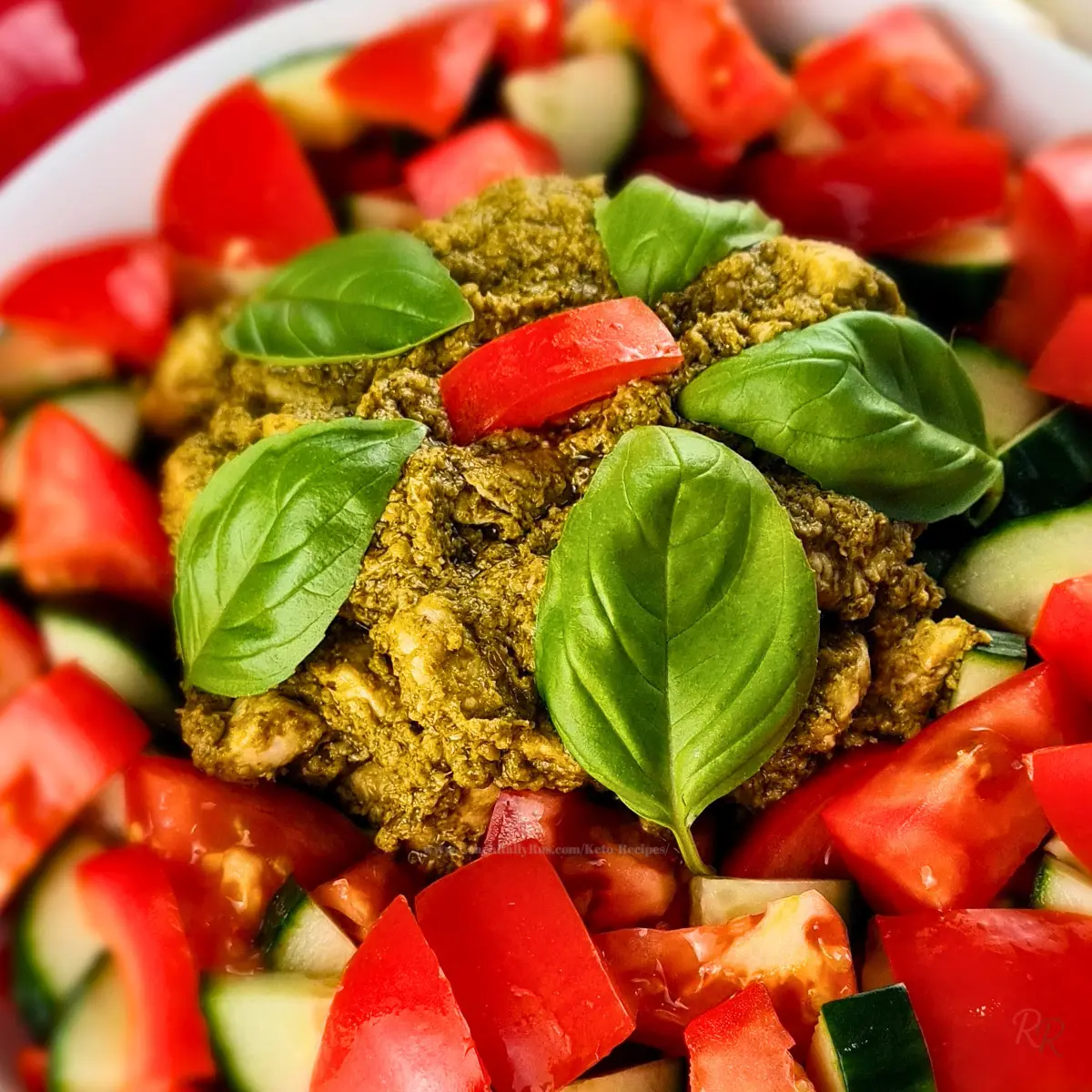 Close-up of pesto chicken on keto chicken salad with chopped tomatoes, cucumber, red bell pepper, and basil leaves