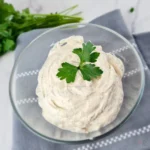 Keto ranch dressing with Greek yogurt in a glass bowl with parsley on top and fresh herbs in the background