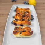 Sliced lemon blueberry cottage cheese bars on a white platter with fresh blueberries and lemon zest on a wooden table.