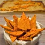 Bowl of nacho cottage cheese chips with bright orange seasoning, showing crisp triangular chips stacked in a parchment-lined bowl with the baked sheet pan in the background.
