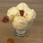 Three creamy scoops of maple brown butter pecan cottage cheese ice cream in a glass coupe topped with a pecan half, with scattered pecans on a wooden surface.