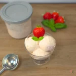 Ninja Creami strawberry basil cottage cheese ice cream in a glass sundae bowl with the frozen pint container and ice cream scoop alongside, garnished with fresh strawberry and basil