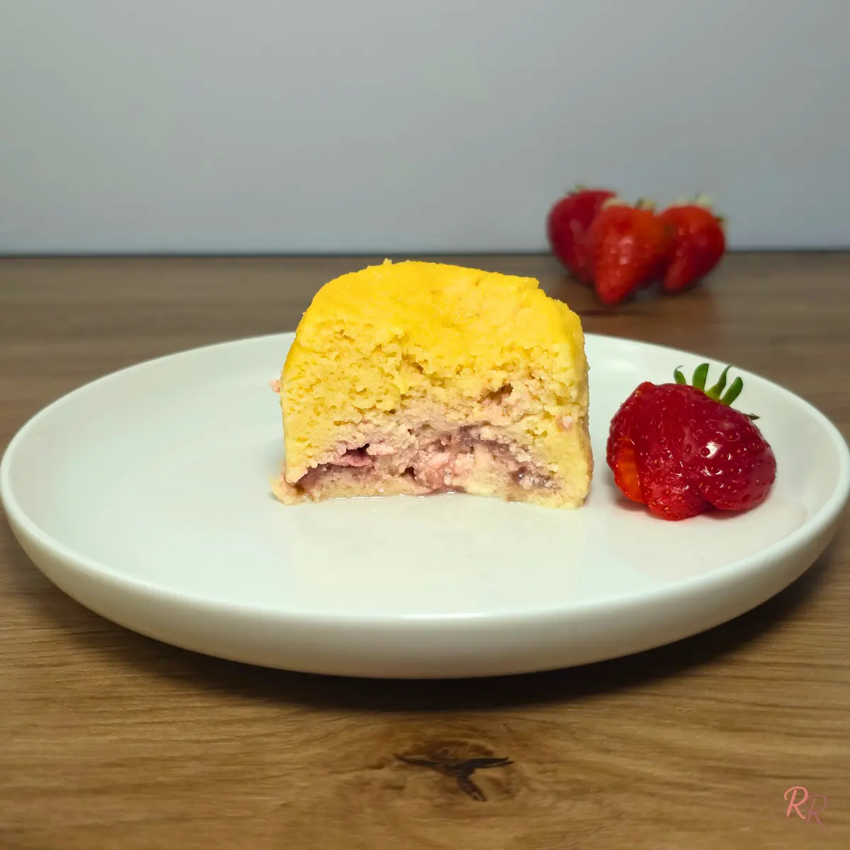 Strawberry cheesecake cottage cheese mug cake cut in half on a white plate showing the cottage cheese cake and cream cheese strawberry center with fresh strawberries