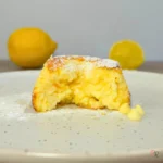 Close-up of a lemon white chocolate cottage cheese mug cake cut open to reveal the moist golden interior, with powdered sugar on top and white chocolate chips beside it on a speckled white plate