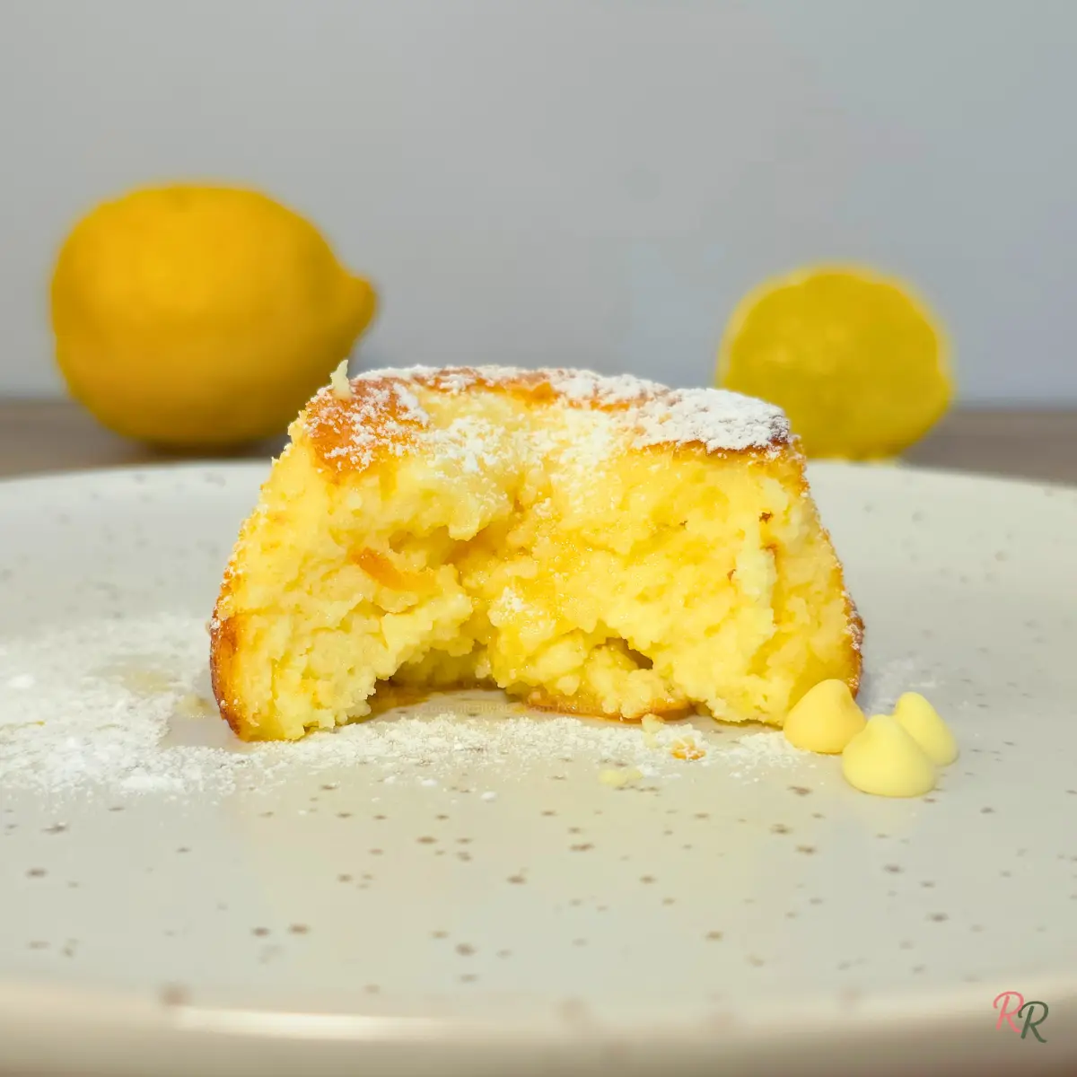 Close-up of a lemon white chocolate cottage cheese mug cake cut open to reveal the moist golden interior, with powdered sugar on top and white chocolate chips beside it on a speckled white plate