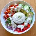 White bowl of Mediterranean salad on a bamboo board with halved cherry tomatoes, sliced cucumber, red bell pepper strips, sliced red onion, chopped parsley and crumbled feta cheese, creamy dressing drizzled over, and a small glass dish of thick whipped feta cottage cheese dressing in the centre