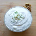 Glass dish of thick whipped feta cottage cheese dressing on a bamboo board, swirled smooth and speckled with herbs, topped with crumbled feta cheese and green herb flecks, with walnut halves beside it