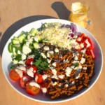 Taco ground beef salad in a white bowl with cilantro lime cottage cheese dressing drizzled over the beef, cucumber, cherry tomatoes, cheddar, red onion, bell pepper and pepitas