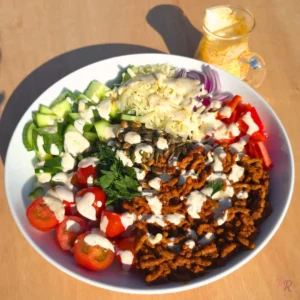 Taco ground beef salad in a white bowl with cilantro lime cottage cheese dressing drizzled over the beef, cucumber, cherry tomatoes, cheddar, red onion, bell pepper and pepitas