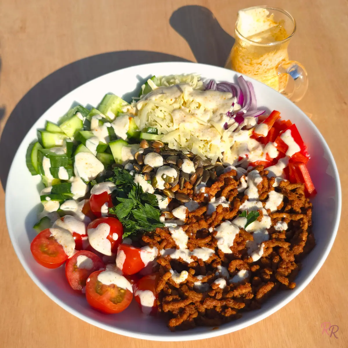 Taco ground beef salad in a white bowl with cilantro lime cottage cheese dressing drizzled over the beef, cucumber, cherry tomatoes, cheddar, red onion, bell pepper and pepitas